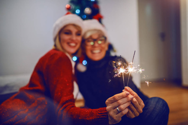 Happy senior woman and her daughter celebrating new year. Both having santa hats on heads and holding sparklers. In background is christmas tree. Family values concept. Selective focus on hands. - Valokuva, kuva