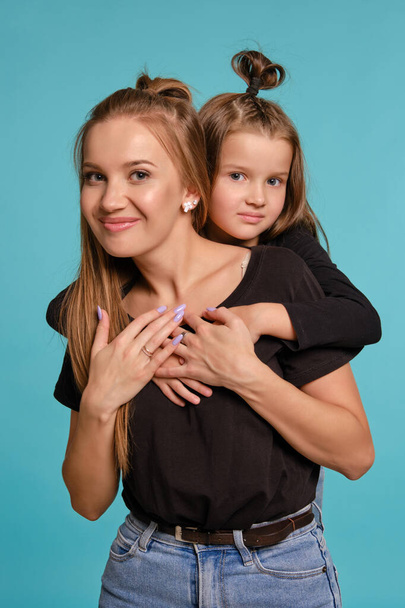 Η μαμά και η κόρη με ένα αστείο χτένισμα, ντυμένες με μαύρα πουκάμισα και μπλε τζιν ποζάρουν σε ένα μπλε φόντο στούντιο. Κοντινό πλάνο.. - Φωτογραφία, εικόνα