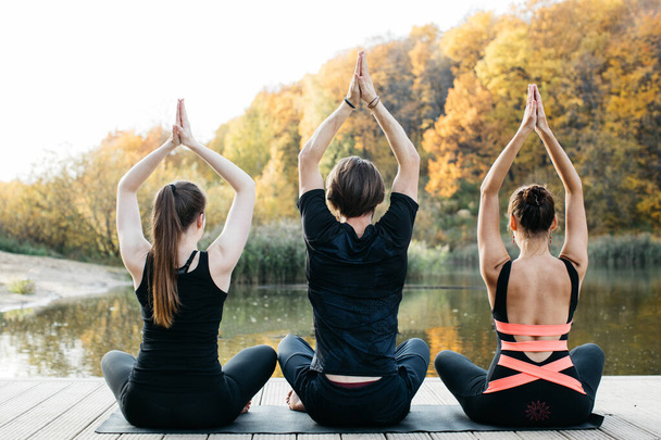 Young women and man doing yoga asana in the nature with the lake view. Evening workout outdoors, sports and healthy lifestyle - 写真・画像