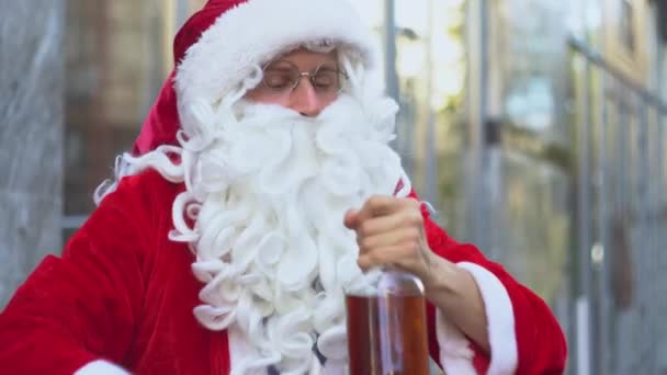 Bad drunk Santa Claus sits near a city office building. Santa Claus drinks alcohol from a bottle on the street - Кадри, відео