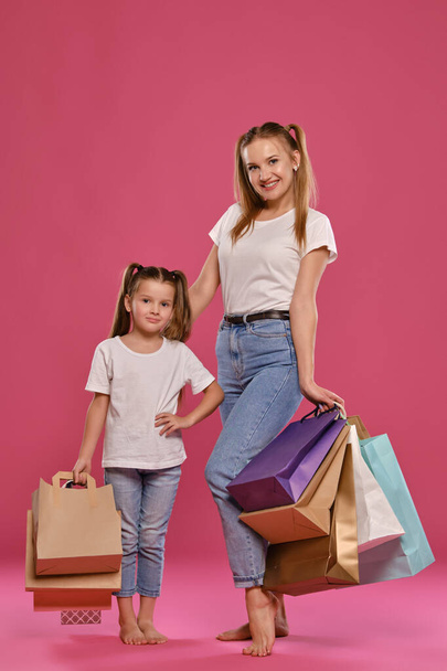 Mom and daughter with ponytails, dressed in white t-shirts and blue jeans are posing against pink background with packages in hands. Full length. - Foto, Imagen