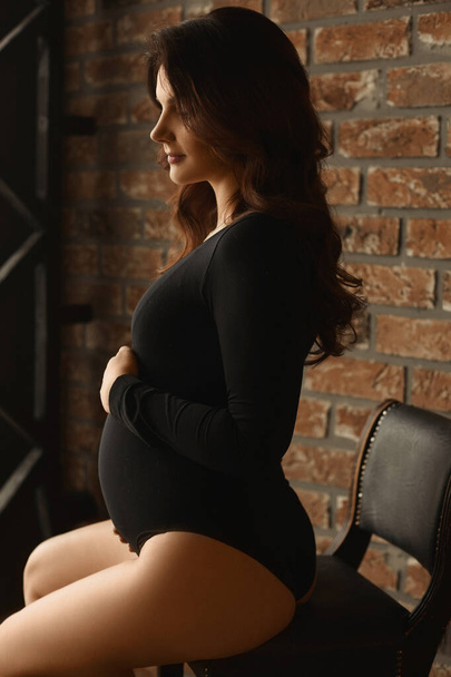 Side view portrait of a beautiful pregnant young woman in black bodysuit sitting in front of a brick wall in interior and holding with love her belly. Last month of pregnancy. Concept of motherhood - Fotó, kép