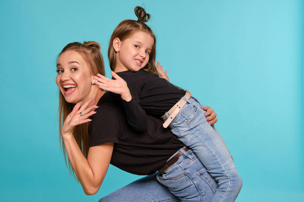 Η μαμά και η κόρη με ένα αστείο χτένισμα, ντυμένες με μαύρα πουκάμισα και μπλε τζιν ποζάρουν σε ένα μπλε φόντο στούντιο. Κοντινό πλάνο.. - Φωτογραφία, εικόνα