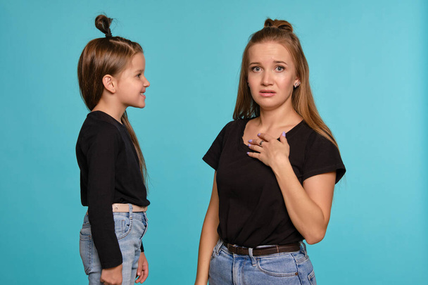 Mom and daughter with a funny hairstyles, dressed in black shirts and blue denim jeans are posing against a blue studio background. Close-up shot. - Photo, image
