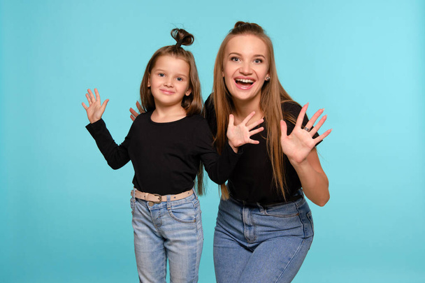 Mutter und Tochter mit lustigen Frisuren posieren in schwarzen Hemden und blauen Jeans vor blauem Studiohintergrund. Nahaufnahme. - Foto, Bild