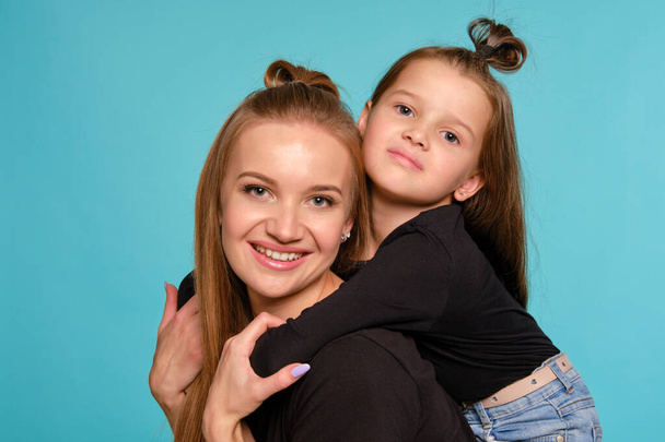 Mutter und Tochter mit lustigen Frisuren posieren in schwarzen Hemden und blauen Jeans vor blauem Studiohintergrund. Nahaufnahme. - Foto, Bild