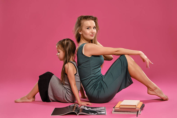 Maman et sa fille vêtues de robes grises posent assises sur un plancher avec un magazine et des livres sur fond de studio rose. Gros plan moyen. - Photo, image