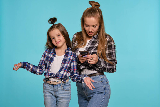 Mom and daughter dressed in checkered shirts and blue denim jeans are using smartphone while posing against a blue studio background. Close-up shot. - Photo, Image