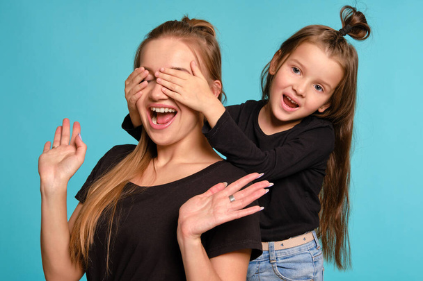 Mom and daughter with a funny hairstyles, dressed in black shirts and blue denim jeans are posing against a blue studio background. Close-up shot. - Valokuva, kuva
