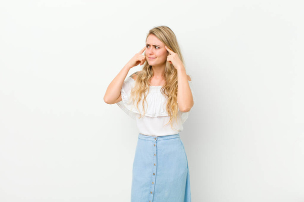 young blonde woman looking concentrated and thinking hard on an idea, imagining a solution to a challenge or problem against white wall - Photo, Image