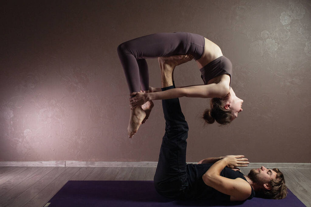 Young sporty man and girl practicing yoga, meditating in yoga pose, working out, wearing sportswear, indoor full length, brown yoga studio - Valokuva, kuva