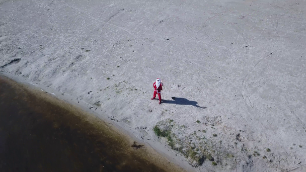 Babbo Natale si trova con una borsa regalo sulla riva sabbiosa del lago. Babbo Natale saluta la telecamera con la mano. Vista dall'alto
 - Filmati, video