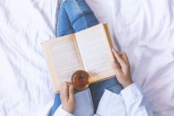 古い本とお茶のカップとベッドの上の女性。手に本とお茶を持っている女性。女性読書本上のベッドで週末の朝 - 写真・画像