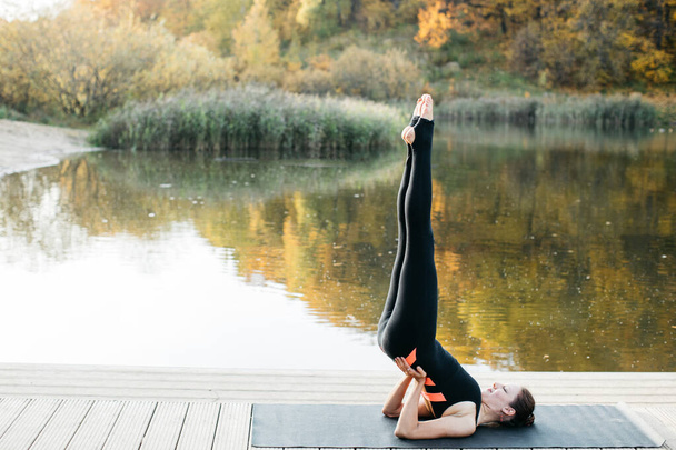 Young woman doing yoga asana in the nature with the lake view. Evening workout outdoors, sports and healthy lifestyle - Photo, Image