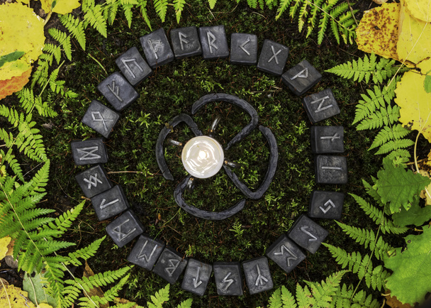 A circle of 24 Scandinavian runes on the background of moss and leaves in the autumn forest. In the center of the circle is a forged candleholder with a candle. - Photo, Image