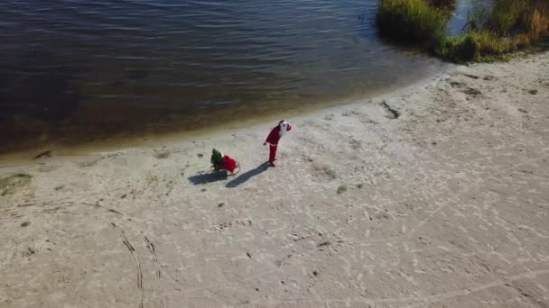 Santa Claus pulls a sleigh with a bag of gifts and a Christmas tree along the sandy shore of the lake. Top view - Footage, Video