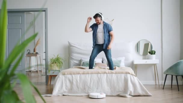Man in headphones dancing on bed while robotic vacuum cleaner vacuuming floor - Filmagem, Vídeo