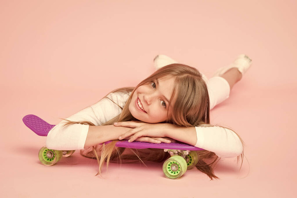 Spreading happy vibrations with the penny board. Small girl skater relaxing at penny board deck on pink background. Little child smiling with violet penny board. Happy hipster with penny skateboard - Foto, afbeelding