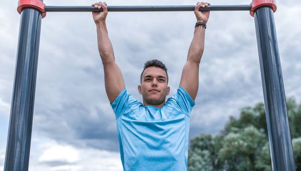 male athlete pulls himself up on bar. In summer spring in city. Active lifestyle, workout, fitness in fresh air. Motivation for sports. Blue t-shirt, cloud background. - Fotografie, Obrázek