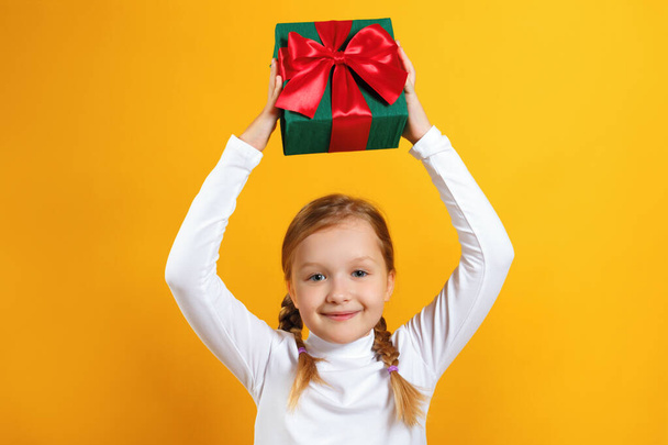 Cute happy little girl holding a box with a gift over her head. A child with pigtails in a white turtleneck on a yellow background - Fotó, kép