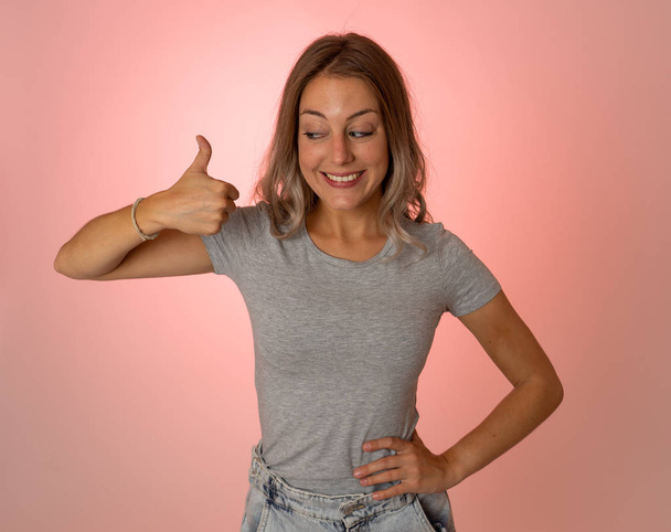 Portrait of beautiful young woman showing thumbs up sign feeling cheerful, happy and satisfied. Young happy student woman making thumb up in joyful approval gesture isolated on pink background. - 写真・画像