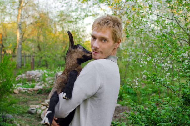 tipo con una pequeña cabra en sus manos en el jardín de primavera
 - Foto, imagen