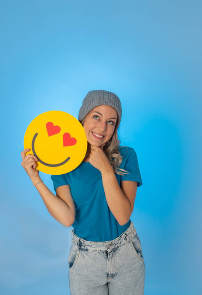 Attractive young woman holding a Hearts Eyes or In Love face emoji feeling happy and excited sharing it in social media. Facial expressions, social network notification icon and technology concept. - Photo, image