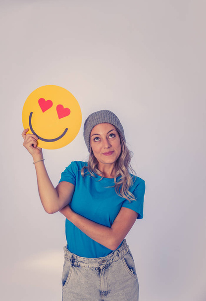 Attractive young woman holding a Hearts Eyes or In Love face emoji feeling happy and excited sharing it in social media. Facial expressions, social network notification icon and technology concept. - Фото, изображение