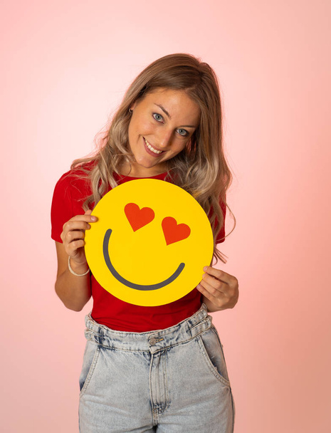 Attractive young woman holding a Hearts Eyes or In Love face emoji feeling happy and excited sharing it in social media. Facial expressions, social network notification icon and technology concept. - Photo, Image