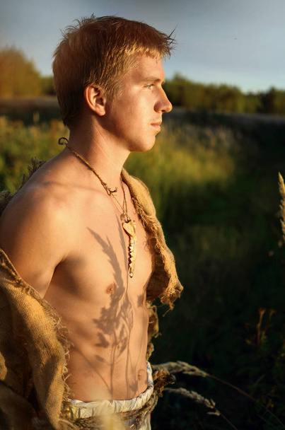 νεαρός άνδρας με ένα κοστούμι από παλιά λινάτσα με ένα οστεοφυλάκιο στο γυμνό στήθος του στέκεται σε ένα χωράφι και κοιτάζει μέσα στην απόσταση - Φωτογραφία, εικόνα
