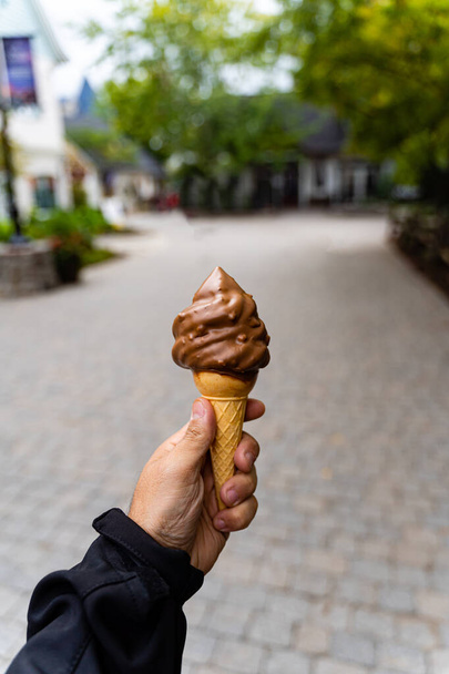 Man holding dipped chocolate ice cream cone in hand, very blurred background, small depth of field. Dark background. - Photo, Image