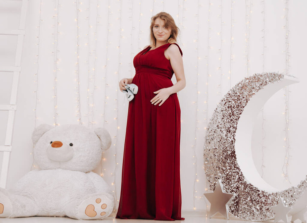 Jeune belle femme enceinte élégante en robe de soirée longue rouge sur fond blanc de la chambre d'enfant
 - Photo, image