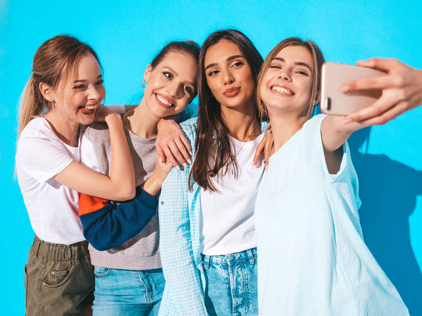 Four young smiling hipster women in summer clothes.Girls taking selfie self portrait photos on smartphone.Models posing near blue wall in studio,Female showing positive face emotions - Zdjęcie, obraz