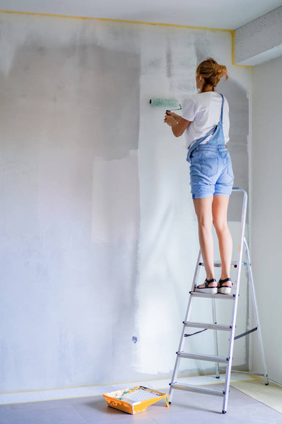 Genç ve güzel bir kadın dairede tamirat yapıyor, duvarları silindirle boyuyor. Molar çalışması. - Fotoğraf, Görsel