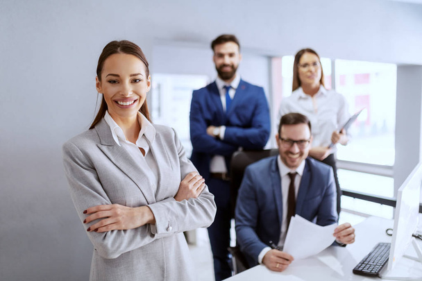 Prachtige lachende zakenvrouw in pak staand in kantoor met handen gekruist. In de achtergrond haar succesvolle team poseren. - Foto, afbeelding