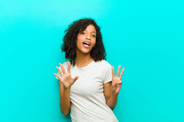 young black woman smiling and looking friendly, showing number eight or eighth with hand forward, counting down against blue wall - Foto, Imagen