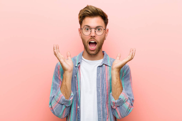 young man feeling shocked and excited, laughing, amazed and happy because of an unexpected surprise against pink background - Foto, Imagem