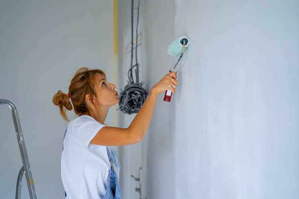 young beautiful woman makes repairs in the apartment, paints the walls with a roller. Molar work. - Фото, изображение