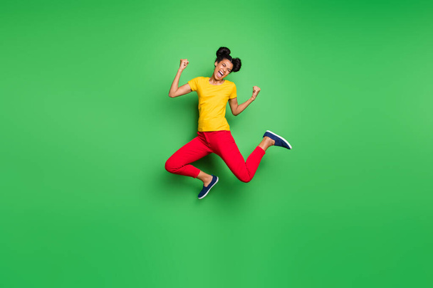 Full size photo of pretty dark skin lady jumping high celebrating exciting victory raising fists wear casual yellow t-shirt red pants isolated green background - Фото, изображение