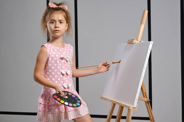 Kleine Künstlerin in rosa Kleid steht hinter Staffelei und malt mit Pinsel auf Leinwand im Kunstatelier mit weißen Wänden. Mittlere Nahaufnahme. - Foto, Bild