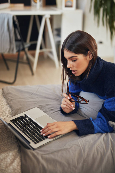 Atrakcyjna biała brunetka w niebiesko-czarnym swetrze leżąca na brzuchu w łóżku i pisząca na laptopie. - Zdjęcie, obraz