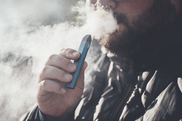 Man smokes new Vape Pod System, inhales and exhales vapor of electronic cigarette, vaping concept - Photo, image