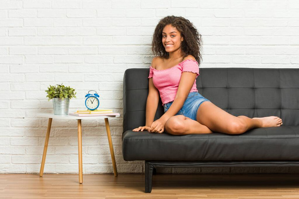 junge afrikanisch-amerikanische Frau sitzt auf dem Sofa und schaut lächelnd zur Seite, fröhlich und angenehm. - Foto, Bild