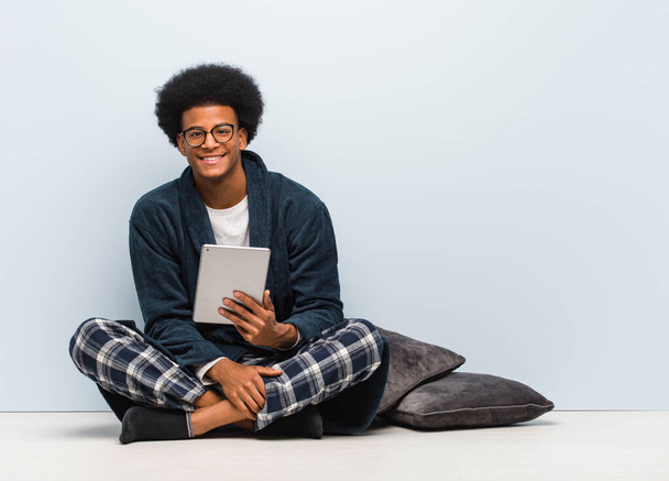 Νεαρός μαύρος κάθεται στο σπίτι του και κρατάει το τάμπλετ του χαρούμενο με ένα μεγάλο χαμόγελο - Φωτογραφία, εικόνα