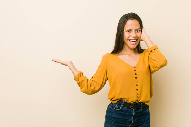 Jonge Latijns-Amerikaanse vrouw tegen een beige achtergrond houdt kopieerruimte op een handpalm, hand op de wang. Verbaasd en verheugd. - Foto, afbeelding