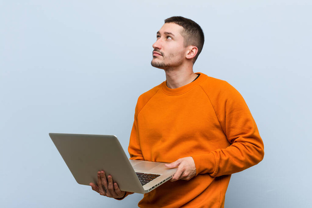 młody biały mężczyzna trzymając laptop uśmiechnięty pewny siebie z skrzyżowanymi ramionami. - Zdjęcie, obraz