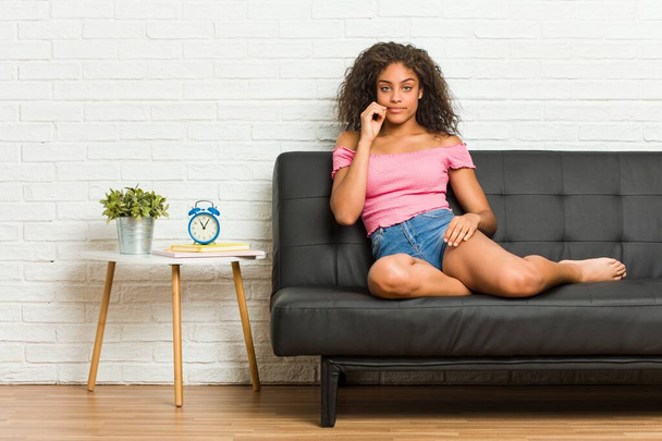Молодая африканская американка, сидящая на диване с пальцами на губах и хранящая секрет
. - Фото, изображение