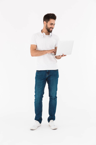 Full length image of pleased young man with headphones over neck smiling and using laptop - Фото, зображення