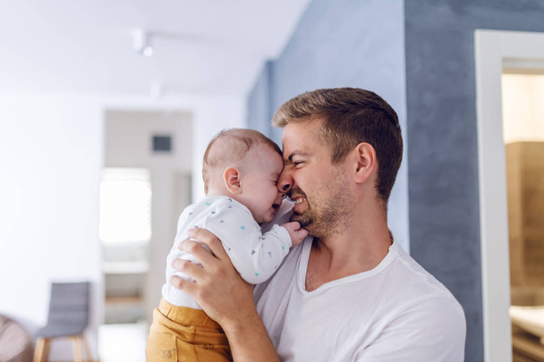 Bonito caucasiano jovem pai segurando seu amoroso filho de 6 meses de idade. O bebé está a brincar com a barba do pai. Interior da sala. Conceito de amor incondicional
. - Foto, Imagem