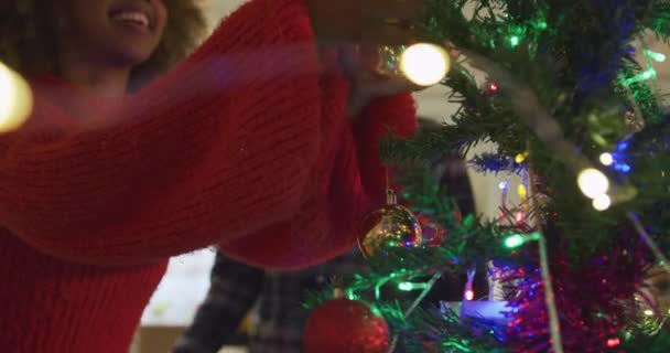 Front view of a smiling young mixed race man and woman hanging a decoration on their Christmas tree at home and embracing beside it - Záběry, video